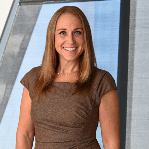 Professional caucasian woman with light brown hair and brown top standing in front of a window