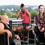 Four disabled people of color with canes and prosthetic legs laugh while chatting. They are on a rooftop deck, in chairs of various height, with greenery and city high-rises in the background.