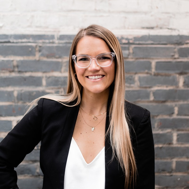 cait Smiling caucasian woman with long blonde hair wearing glasses, a black jacket, and white shirt standing in front of a brick wall