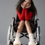 Young woman of color in red sweater and white boots sitting in wheelchair with her chin in her hands