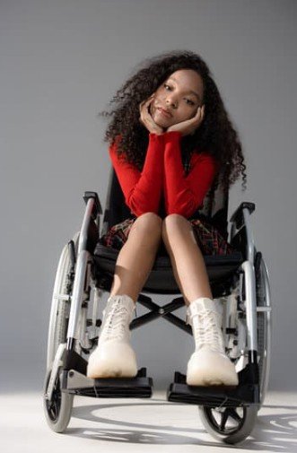 Young woman of color in red sweater and white boots sitting in wheelchair with her chin in her hands