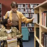 Two students, one standing and one sitting in a wheelchair, choosing books in a library