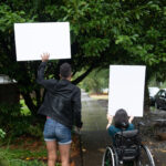 A Black non-binary person with a cane and a South Asian person in a wheelchair block a neighborhood street while holding up cardboard signs. The photo is shot from behind them.