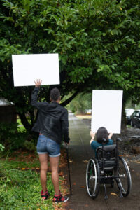 A Black non-binary person with a cane and a South Asian person in a wheelchair block a neighborhood street while holding up cardboard signs advocating for accessibility and inclusion. The photo is shot from behind them.
