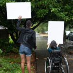 A Black non-binary person with a cane and a South Asian person in a wheelchair block a neighborhood street while holding up cardboard signs. The photo is shot from behind them.