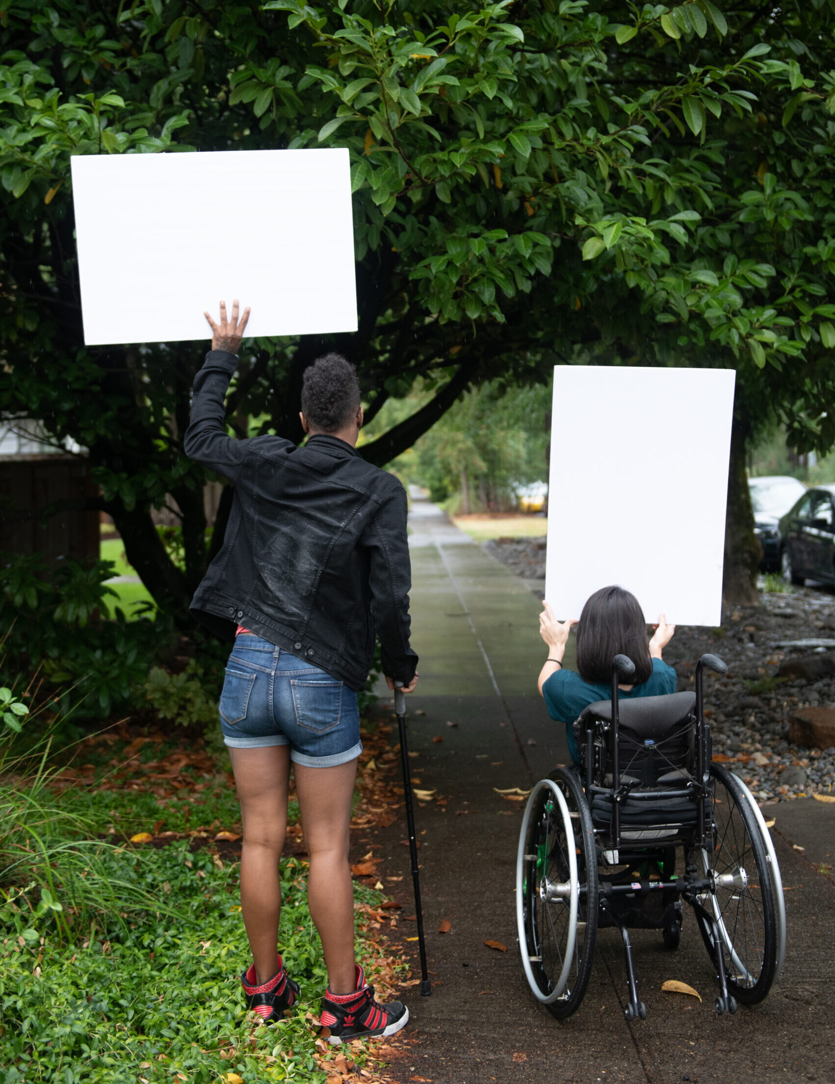A Black non-binary person with a cane and a South Asian person in a wheelchair block a neighborhood street while holding up cardboard signs. The photo is shot from behind them.